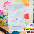 White package with 'Wildflower & Ember' branding on a table with art supplies and floral background